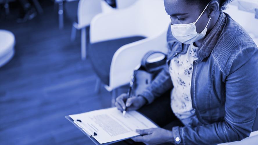 A black woman wearing a medical mask fills out a form in a waiting room. Photo by Drazen Zigic/Getty Images