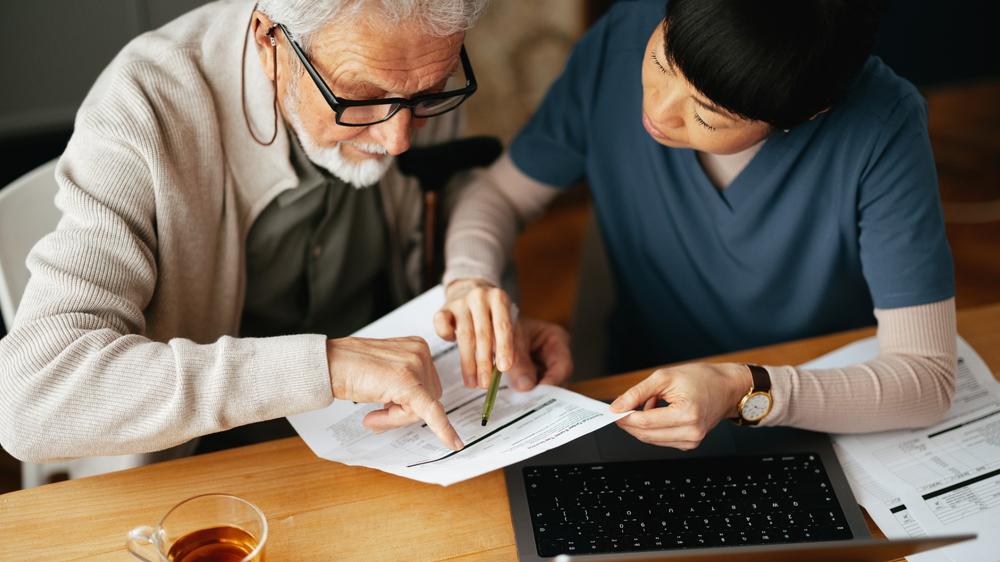 Health care staff person and an elderly man looking at a bill in front of a laptop in the man's home
